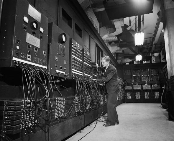 A black and white photo of ENIAC, an antiquated computer taking up an entire room as a man leans into to look at it.